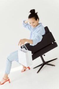 Smiling woman with hair bun holding a shopping bag, sitting on a black chair against a white background.